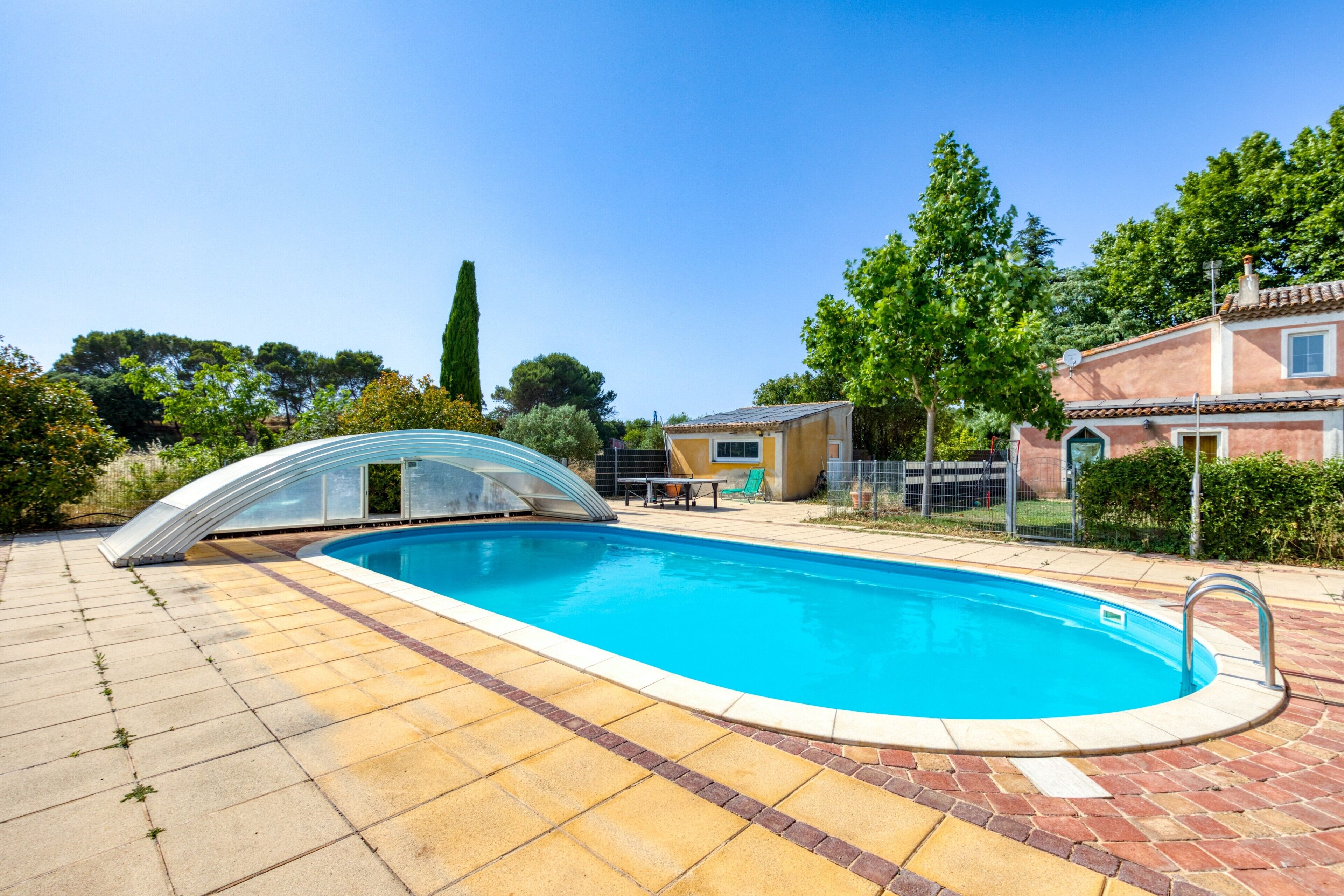 A swimming pool with a glass enclosure, surrounded by a brick patio, and adjacent to a building with a tiled roof.