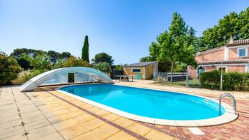A swimming pool with a glass enclosure, surrounded by a brick patio, and adjacent to a building with a tiled roof.