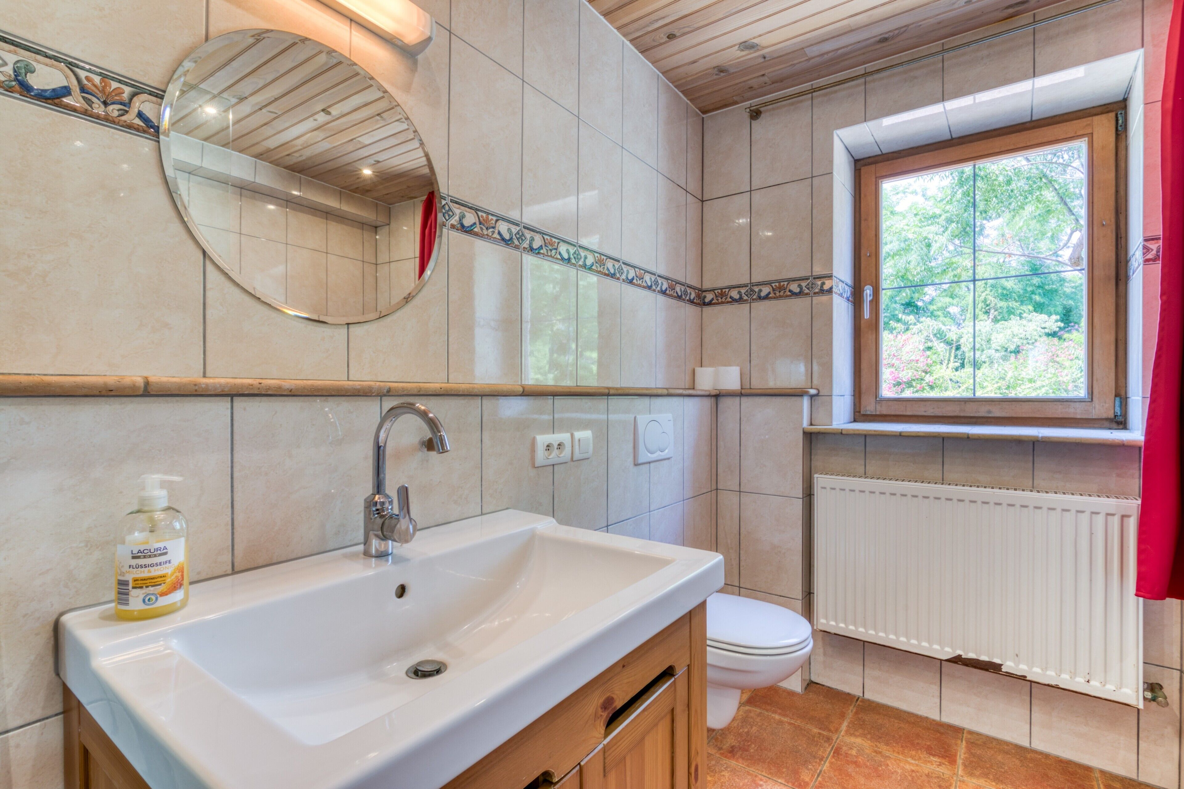 A bathroom with a sink, mirror, toilet, and window with a view of greenery.