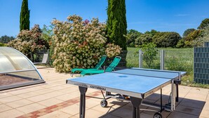 Een overdekt buitengebied met een tafeltennistafel, groene lounge fauteuils en een kas.