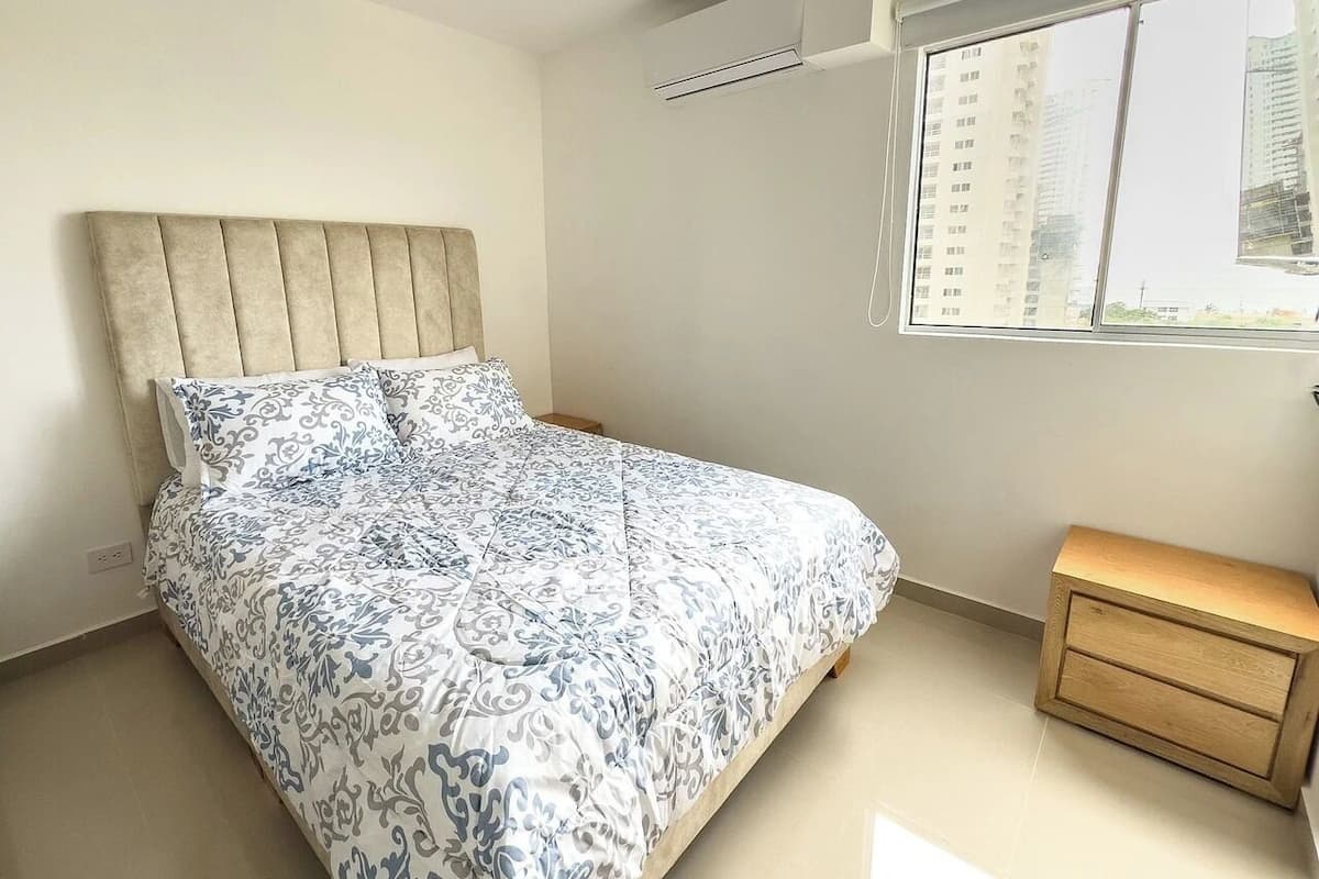 A bedroom with a bed, a bedside table, and a window with a view of buildings.