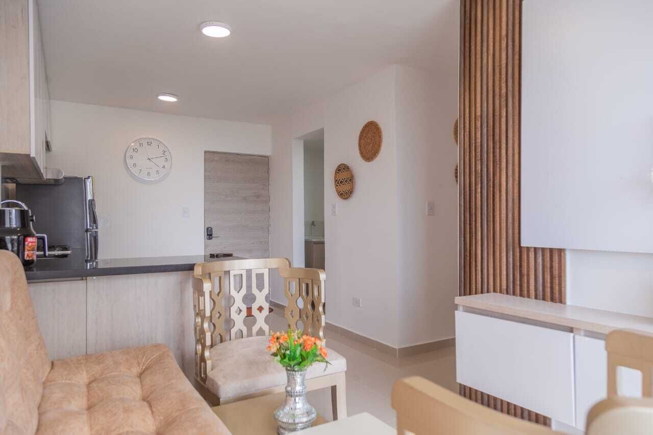 Una sala de estar moderna con sofá, una mesa de comedor con sillas, una zona de cocina con refrigerador y máquina de café, y un reloj decorativo en la pared.