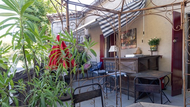 Un patio con una pérgola de metal, mobiliario exterior, plantas en macetas y un edificio con toldo a rayas.