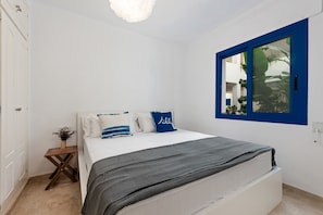 A bedroom with a bed, a small wooden side table, a potted plant, and a window with a blue frame.