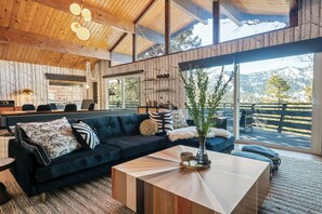 A modern living room with a dark blue sectional sofa, a wooden coffee table, and a large window offering a view of a wooden deck and mountains. - Ski View Chalet (Big Bear Lake)