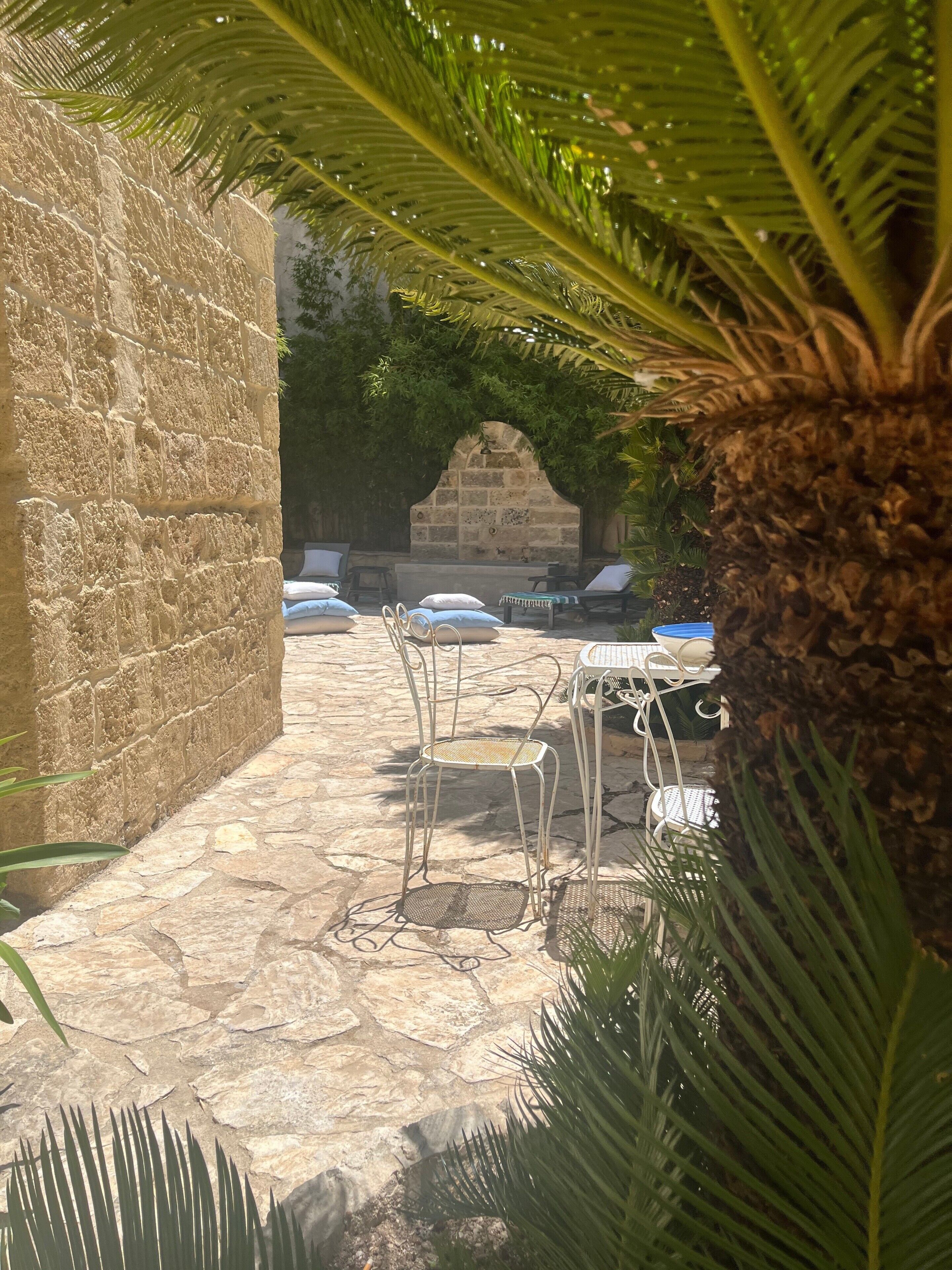 Une terrasse dallée de pierres, avec des espaces de détente en plein air, comprenant des chaises et des tables, entourée de verdure et d’un mur en pierre.