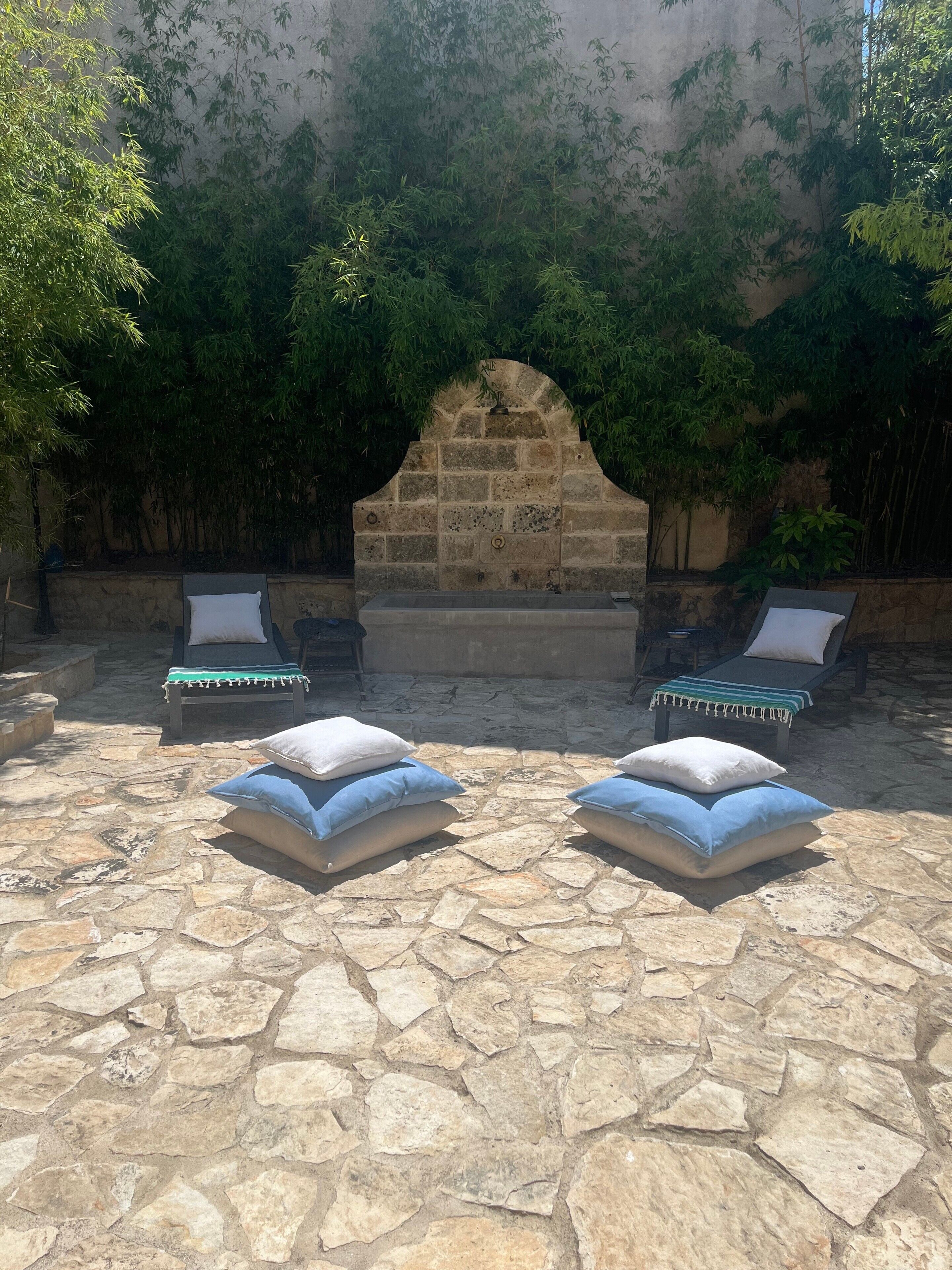 Une terrasse en pierre avec deux chaises longues et des coussins, une structure en pierre en forme d’arche et des feuillages de bambou.