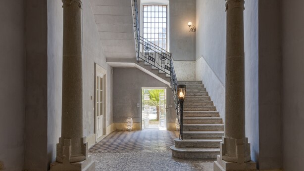 Um hall imponente com piso de mármore, colunas grandes, escadaria com corrimão de metal e vista para o exterior através de uma grande janela.