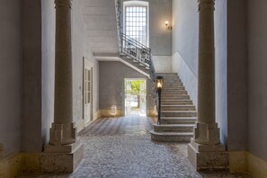 Un amplio pasillo con piso de mármol, grandes columnas, una escalera con barandilla de metal y vistas al exterior a través de un ventanal.