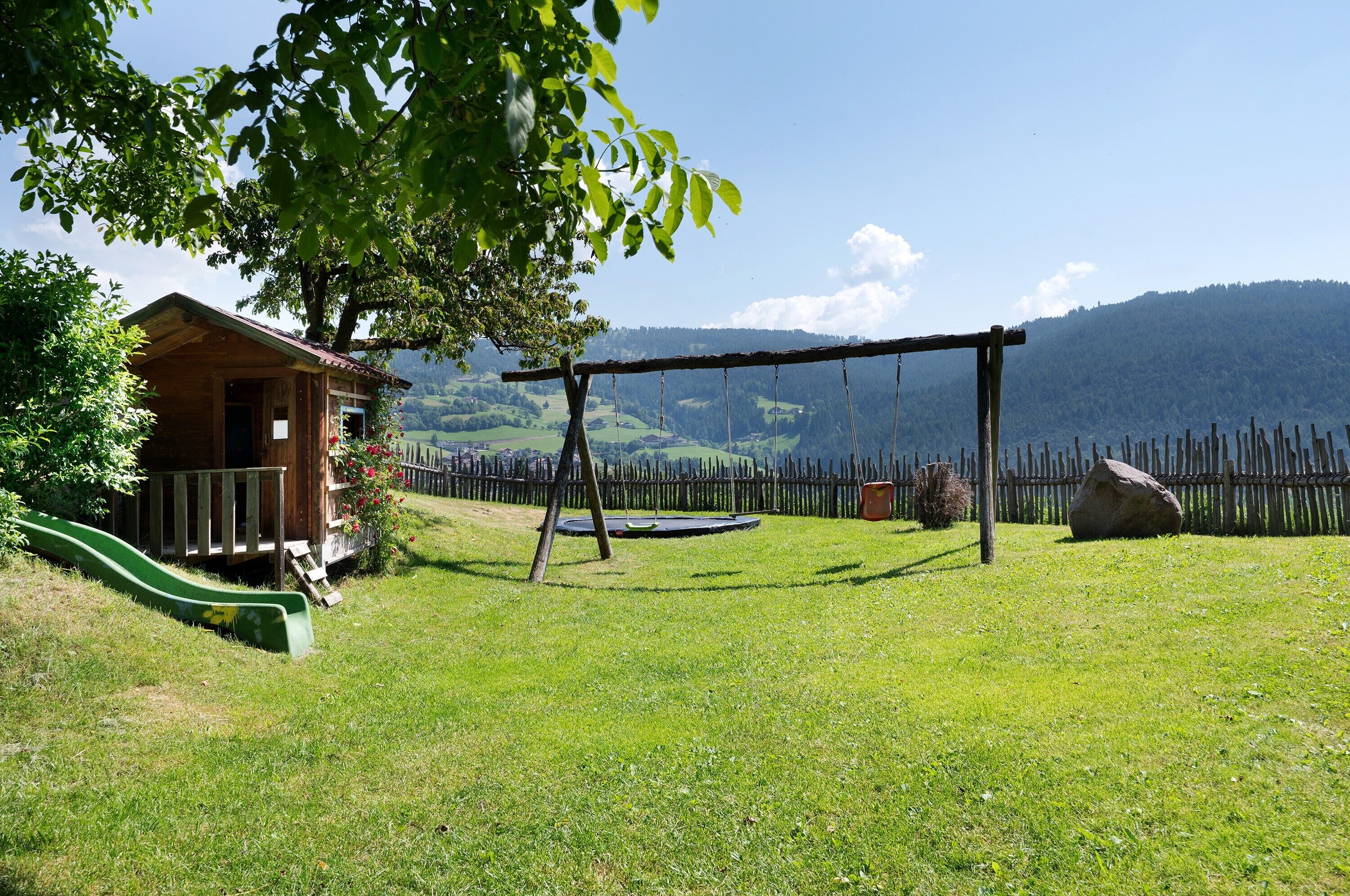 A wooden cabin with a green slide, a swing set, and a fenced yard with a mountain backdrop.