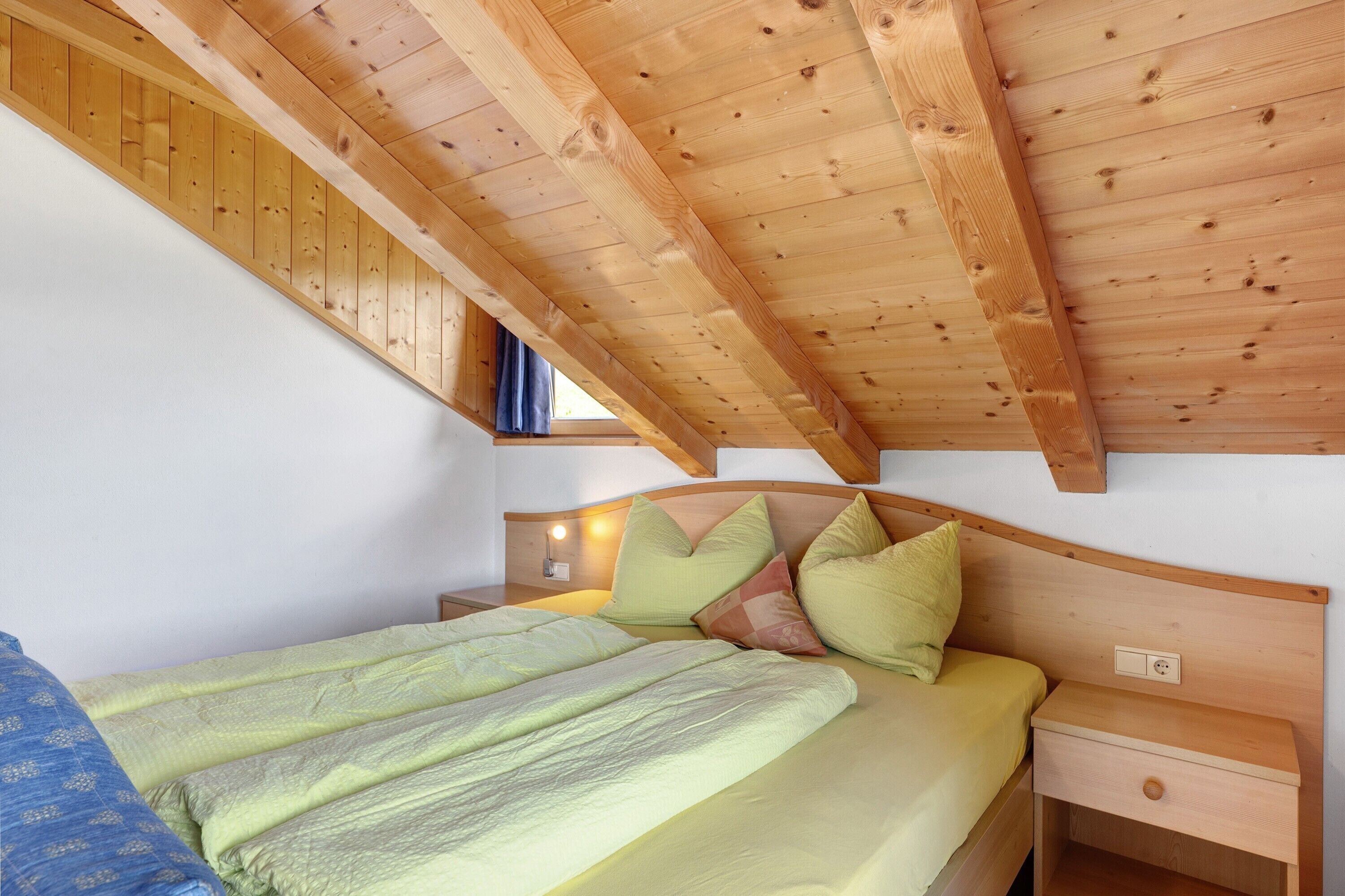 Quarto com teto inclinado, cama com roupa de cama verde, mesa de cabeceira de madeira e tomada na parede.