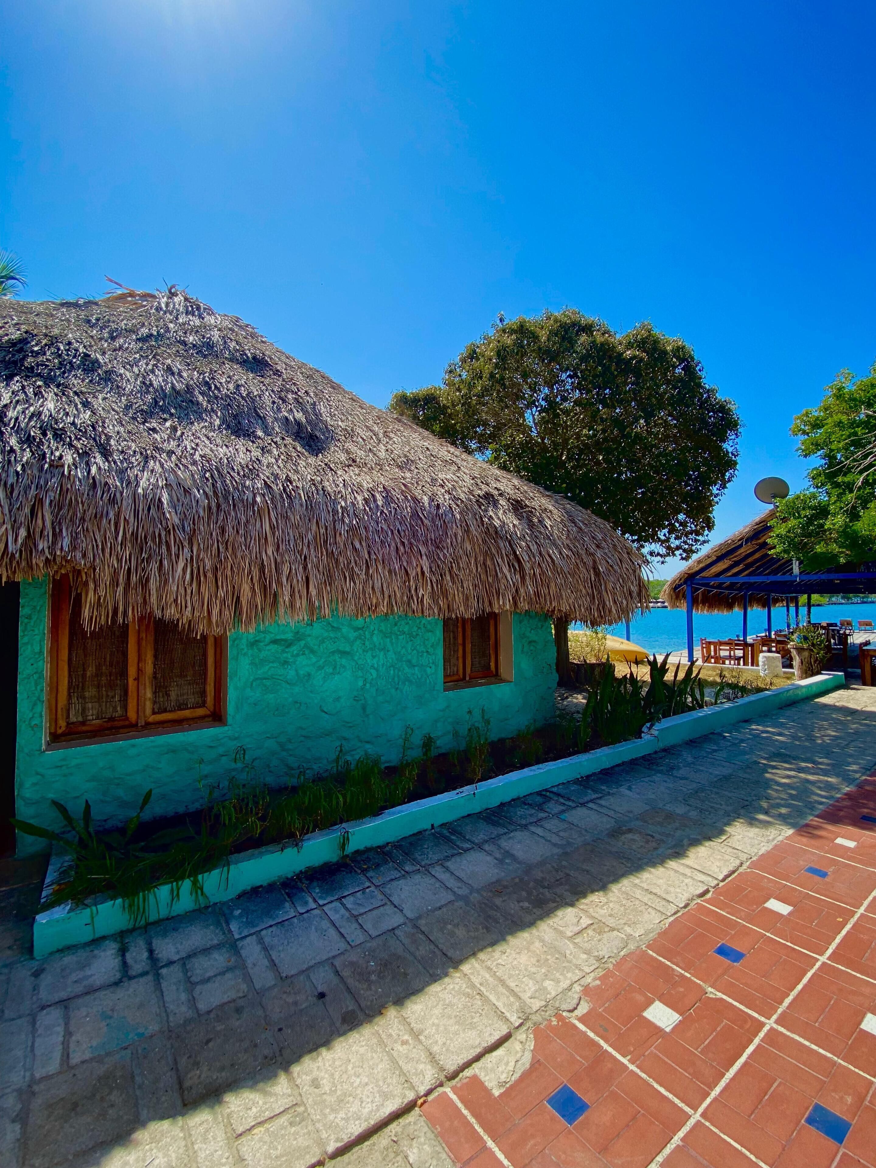 A thatched-roof house with a window and a door, a paved walkway, and a pool area with a blue umbrella.
