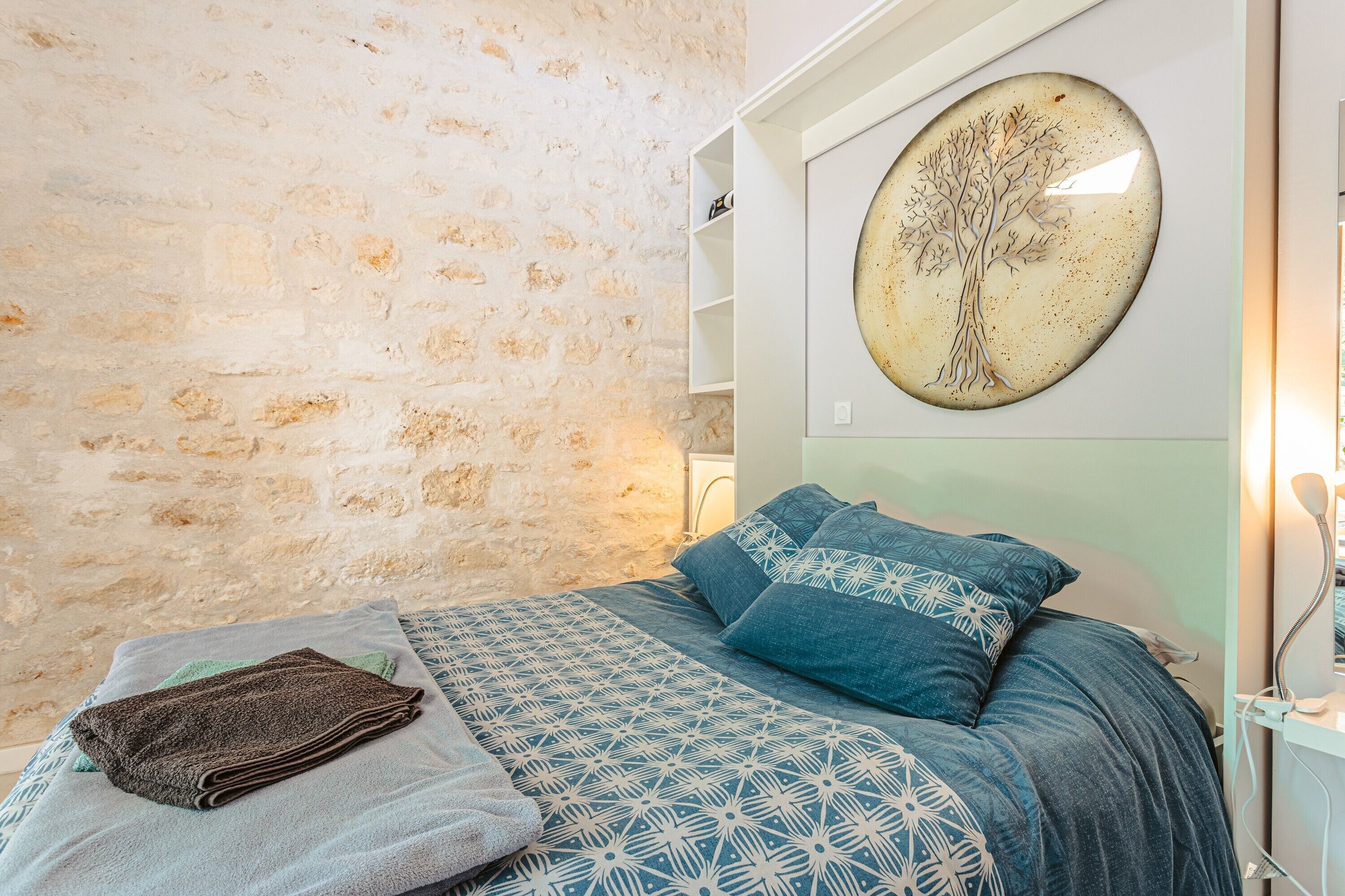 Ein Schlafzimmer mit Steinwand, einem Bett mit gemusterten Betttextilien, einer an der Wand befestigten Baumdarstellung und einer weißen Schrankwand.