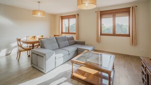 Un salon comprenant un canapé d’angle, une table basse en verre, une table à manger avec des chaises et deux grandes fenêtres avec des rideaux.