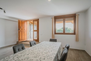 Ein Zimmer mit Esstisch, vier Stühlen, einem Holzschrank, einem Fenster mit Vorhängen und einer Tür, die nach draußen führt.