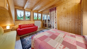 Un dormitorio de cabaña de madera con una cama, un sofá rojo, una mesita de noche con una lámpara y vistas por la ventana.