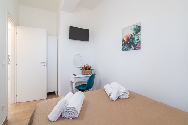 Una habitación de hotel con una cama, un escritorio con silla, un televisor, un espejo en la pared y un cuadro.