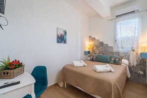 A bedroom with a bed, a chair, a bedside table with a basket of flowers, a wall-mounted air conditioner, and a window with curtains. - Apartment 'Donna Di Denari' with Sea View, Wi-Fi and Air Conditioning (Noto Marina)