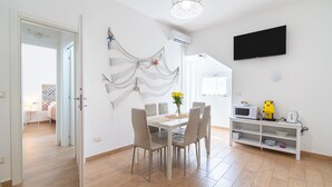 A dining area with a table, chairs, a TV, and a shelf with kitchen appliances. - Apartment 'Donna Di Denari' with Sea View, Wi-Fi and Air Conditioning (Noto Marina)