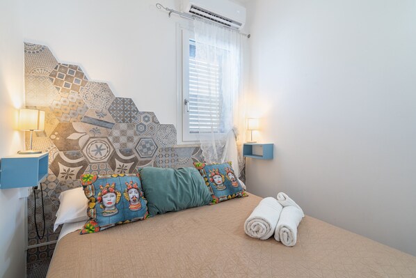 Un dormitorio con un cabecero estampado, dos lámparas de noche, una cama con una toalla enrollada y una ventana con persianas.