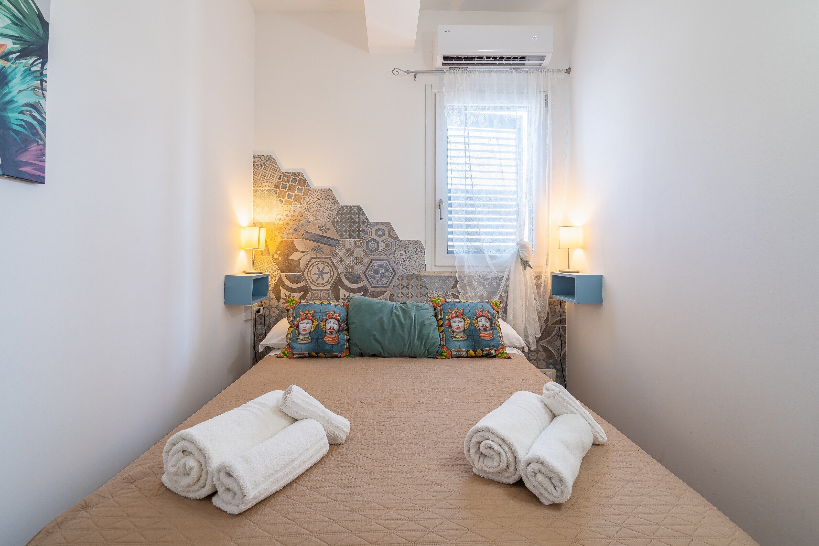 Habitación de hotel con cama, dos lámparas fijadas a la pared, almohadones estampados y una ventana con persianas.