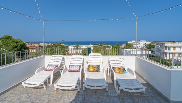 A rooftop terrace with four white lounge chairs, string lights, and a view of the sea. - Apartment 'Donna Di Denari' with Sea View, Wi-Fi and Air Conditioning (Noto Marina)