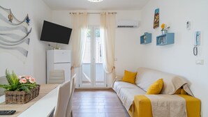 A room with a sofa, a TV, a dining table, and a window with curtains. - Apartment 'Re Di Denari' with Sea View, Wi-Fi and Air Conditioning (Noto Marina)