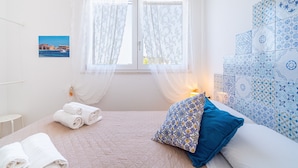 A bedroom with a bed, a bedside table, a window with curtains, a wall with patterned tiles, and a framed picture on the wall. - Apartment 'Re Di Denari' with Sea View, Wi-Fi and Air Conditioning (Noto Marina)