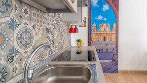 A kitchen with a stainless steel sink, colorful patterned tiles, a small shelf with a bowl of fruit, and a mural of a historic building. - Apartment 'Almàsalo Re Di Coppe' with Private Terrace, Wi-Fi and Air Conditioning (Noto Marina)