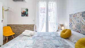 A bedroom with a bed, a yellow chair, a white towel, a window with sheer curtains, and a wall with patterned tiles. - Apartment 'Almàsalo Re Di Coppe' with Private Terrace, Wi-Fi and Air Conditioning (Noto Marina)