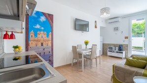 A modern kitchen with a sink, a dining area with a table and chairs, a TV mounted on the wall, and a sofa in the living area.