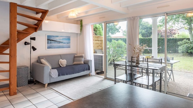 Ein modernes Wohnzimmer mit grauem Sofa, einer Holztreppe, einem Essbereich mit Tisch und Stühlen sowie einer großen Schiebeglastür, die zu einem Garten führt.