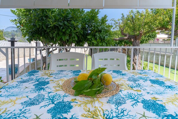 A balcony with a table set for two, a white railing, and a view of a tree and a building in the background.