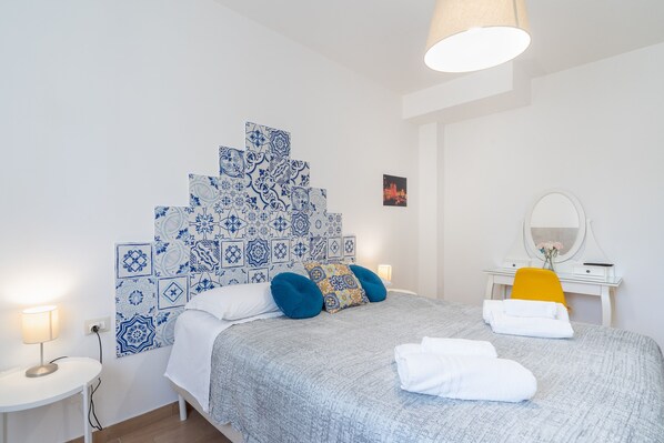 A bedroom with a bed, a bedside table, a desk with a chair, and a wall-mounted mirror.
