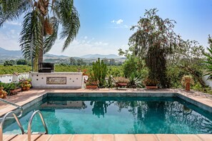 A swimming pool with a view of mountains, surrounded by various plants and a pergola.