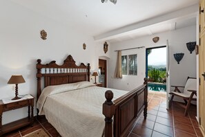 A bedroom with a wooden bed, a nightstand, a lamp, a window with a view of a pool and mountains, and a chair.