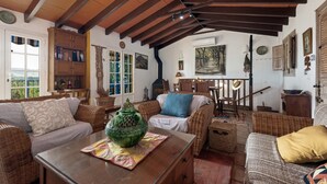 Una sala de estar acogedora con techo de madera alta, muebles de mimbre, una maceta de cerámica verde y un cuadro en la pared.