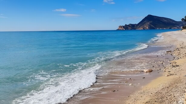 Een strand met helderblauw water, een zandstrand en een berg op de achtergrond.