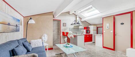 Een moderne keuken met rode kasten, een eethoek met een glazen tafel, en een woonkamer met een blauwe bank.