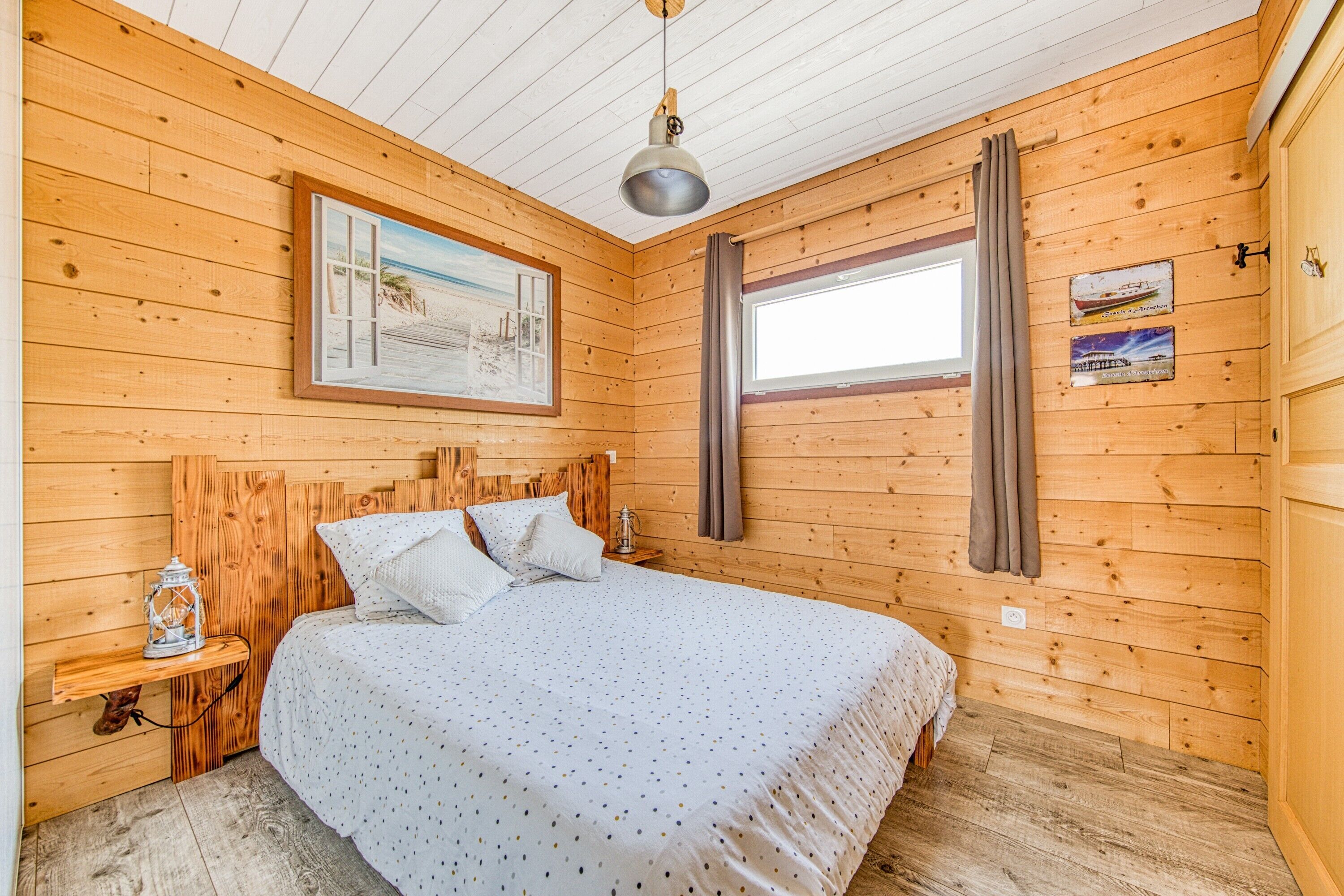 A wooden cabin bedroom with a bed, a nightstand, a hanging lamp, and a window with curtains.