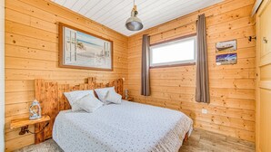 A wooden cabin bedroom with a bed, a nightstand, a hanging lamp, and a window with curtains. - La Gascogne in Andernos-les-Bains – Stylish Apartment for 2, Near Nature & Coast (Andernos-les-Bains)