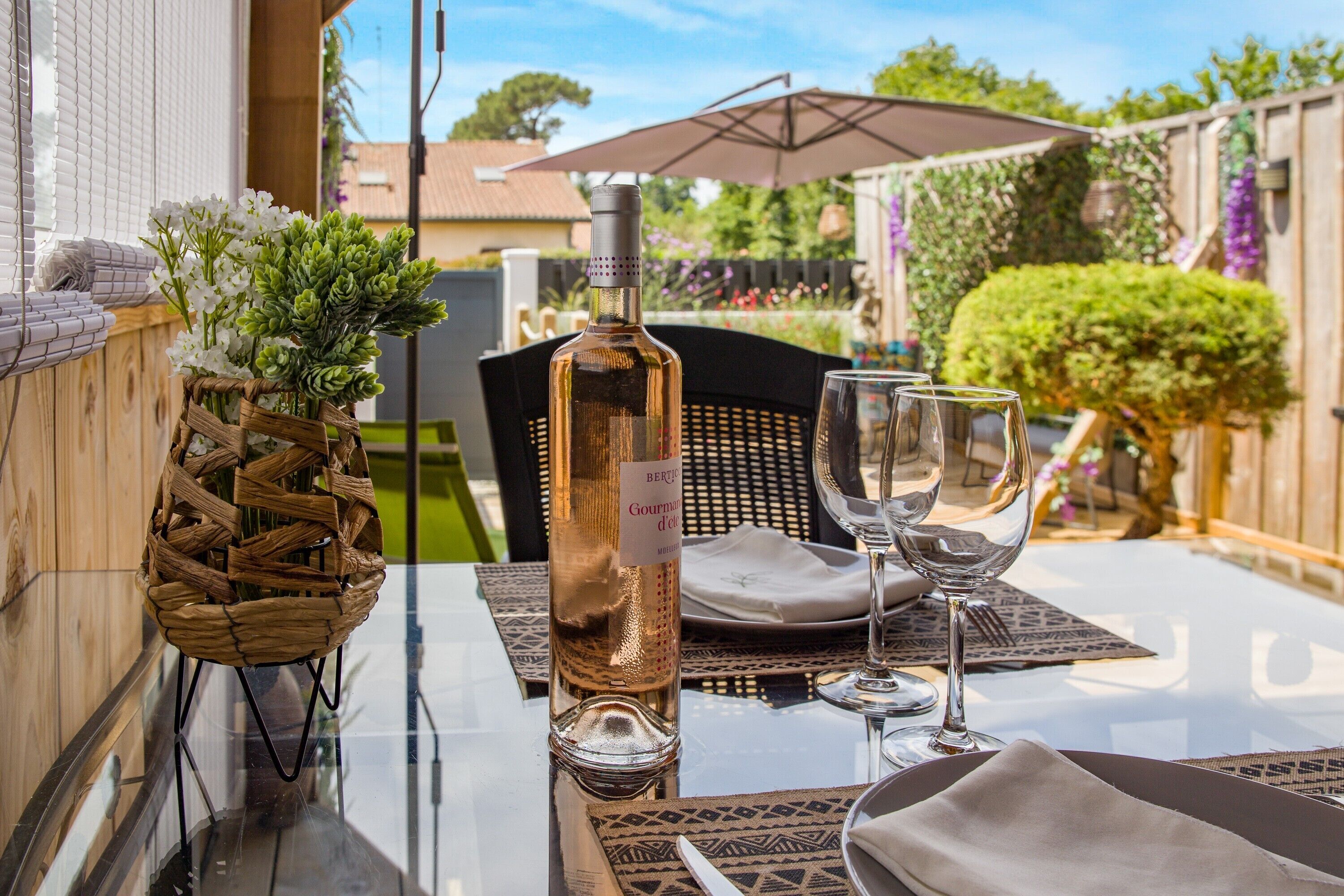 A table set outdoors with a bottle of wine, glasses, and a basket of flowers.