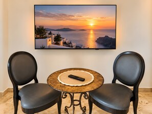 A room with a wall-mounted TV displaying a sunset view, two black chairs, and a small round table with a remote control. - Apartment 'Balneo Luxe' (Ginestas)