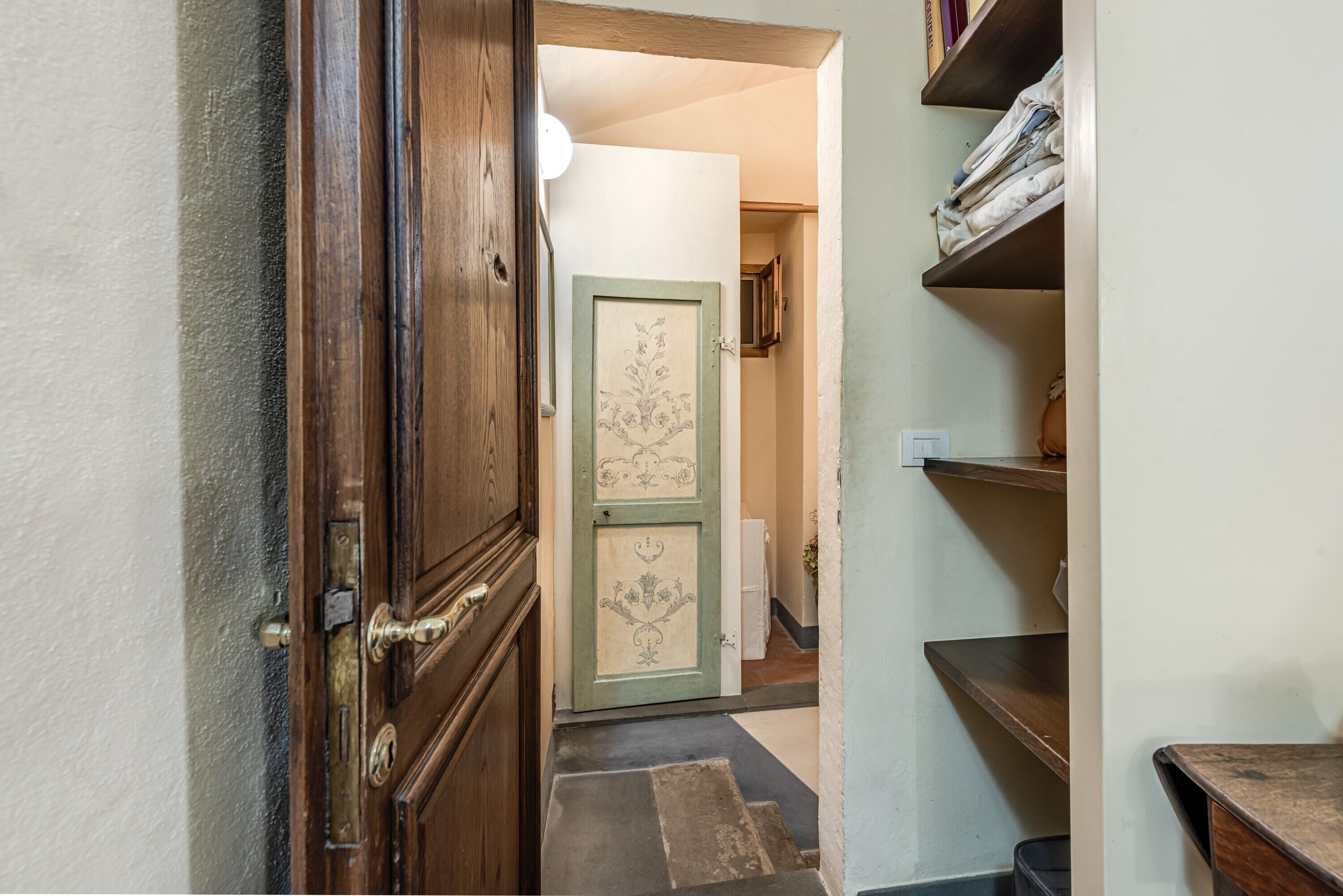 A hallway with a wooden door, a green door with floral patterns, and a shelf with folded clothes.