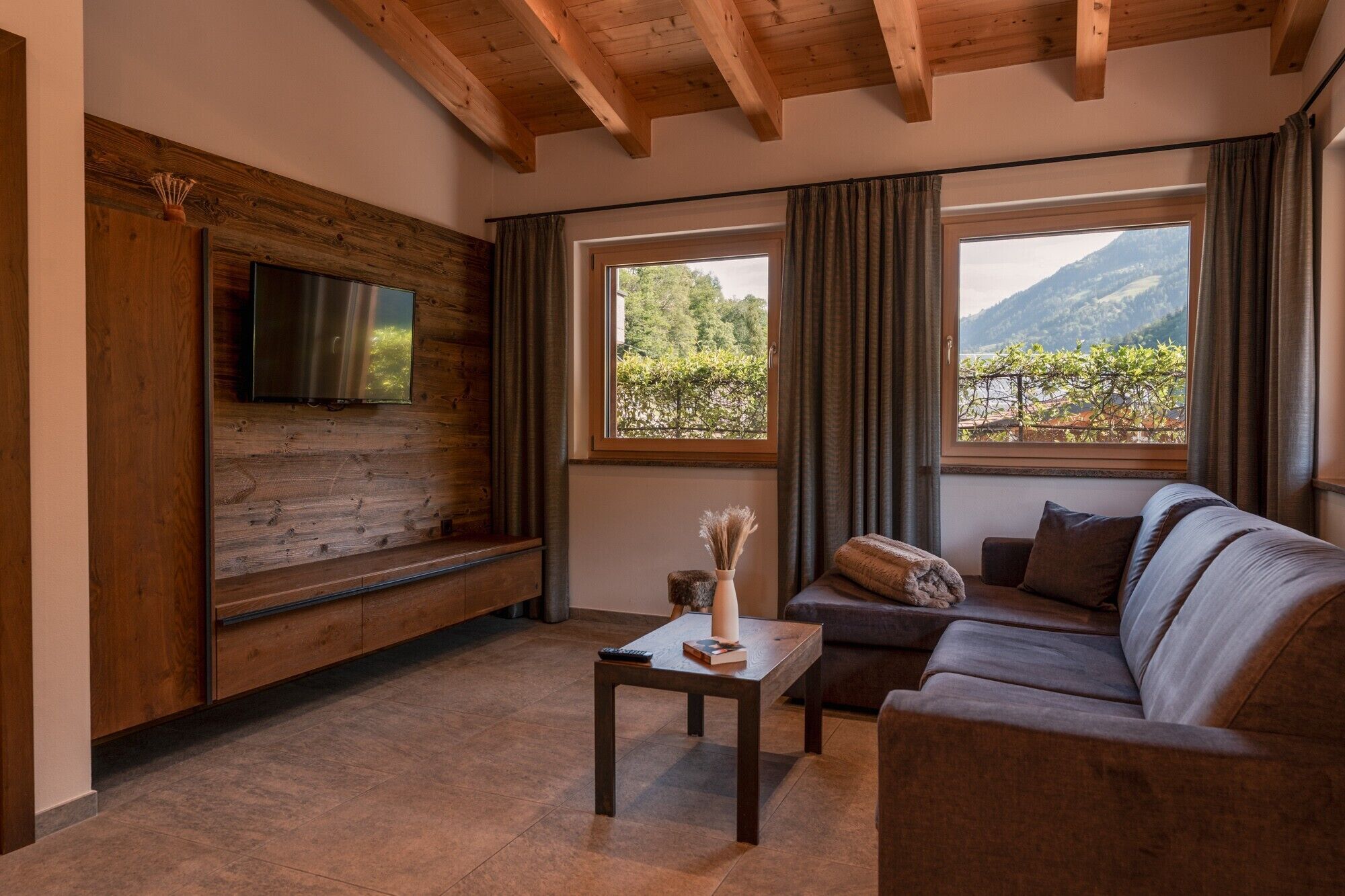A living room with a wooden ceiling, a flat-screen TV mounted on a wooden wall, a sofa, a coffee table, and two windows with curtains.