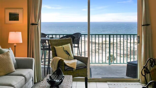 A room with a view of the beach, a wicker chair, a table with a black bowl, and a lamp.