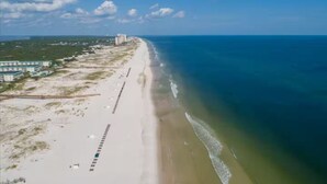 An aerial view of a beach with a long stretch of sand, a few buildings, and a clear blue sky.