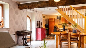 A living room with a piano, a dining area with wooden furniture, a staircase, and a kitchen in the background. - Holiday Home 'De L'alliance' with Private Terrace and Private Garden (Lannion)