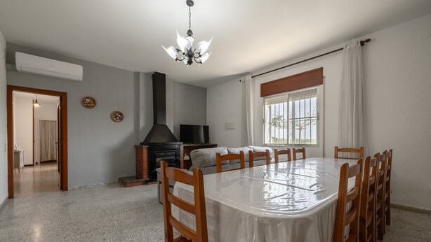 Un comedor con una mesa redonda, sillas de madera, una araña, chimenea y vistas al exterior a través de una ventana con cortinas.