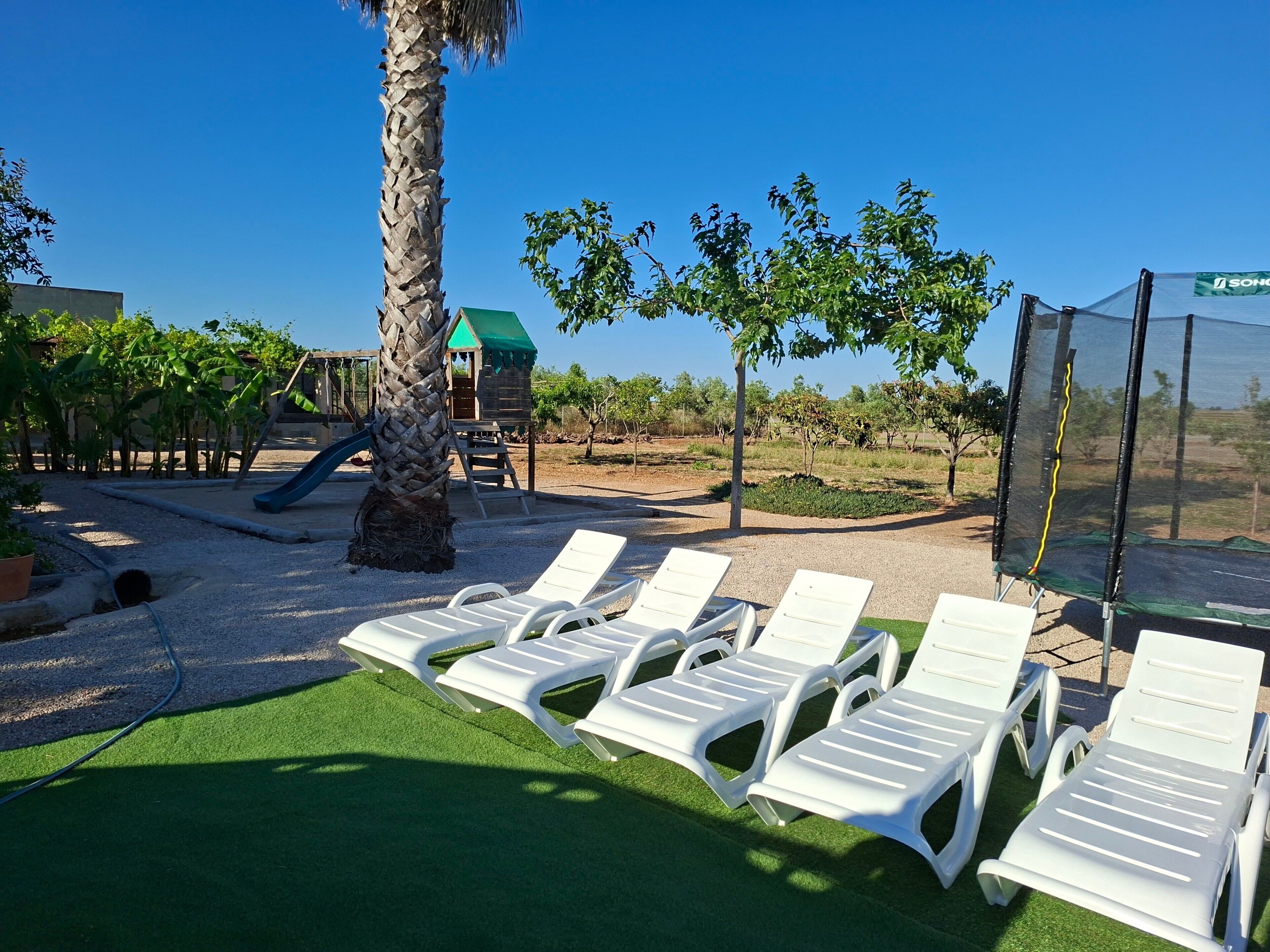 Un parque infantil con tobogán, una palmera y sillas reclinables blancas sobre césped artificial.